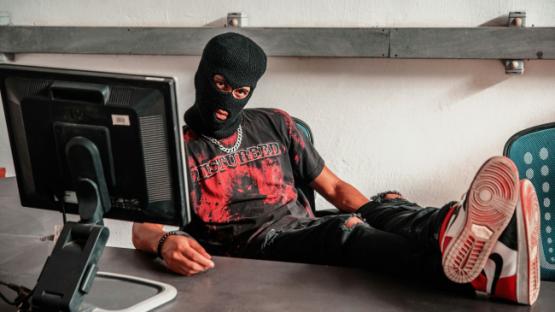 A man wearing a balaclava with his feet on a desk behind a computer screen. His t-shirt reads "DISTURBED".