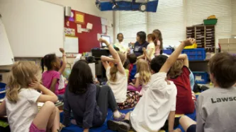 typical classroom scene, where an audience of school children were seated on the floor before a teacher at the front of the room, who was reading an illustrated storybook, during one of the scheduled classroom sessions.