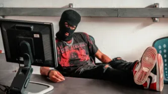 A man wearing a balaclava with his feet on a desk behind a computer screen. His t-shirt reads "DISTURBED".
